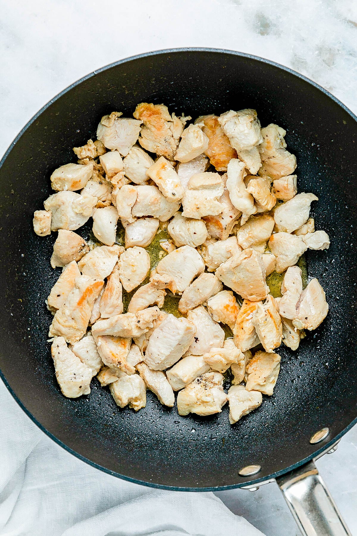 Chunks of chicken cooking in a black frying pan on a light surface.