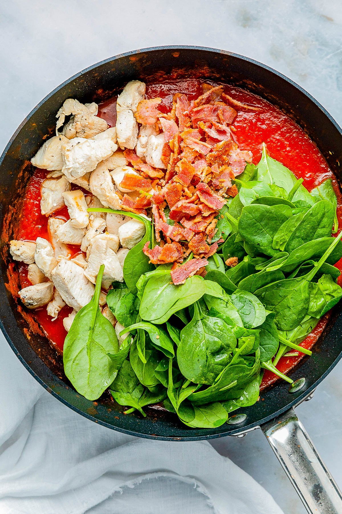 A skillet with cooked chicken, crispy bacon, fresh spinach, and tomato sauce on a light surface.