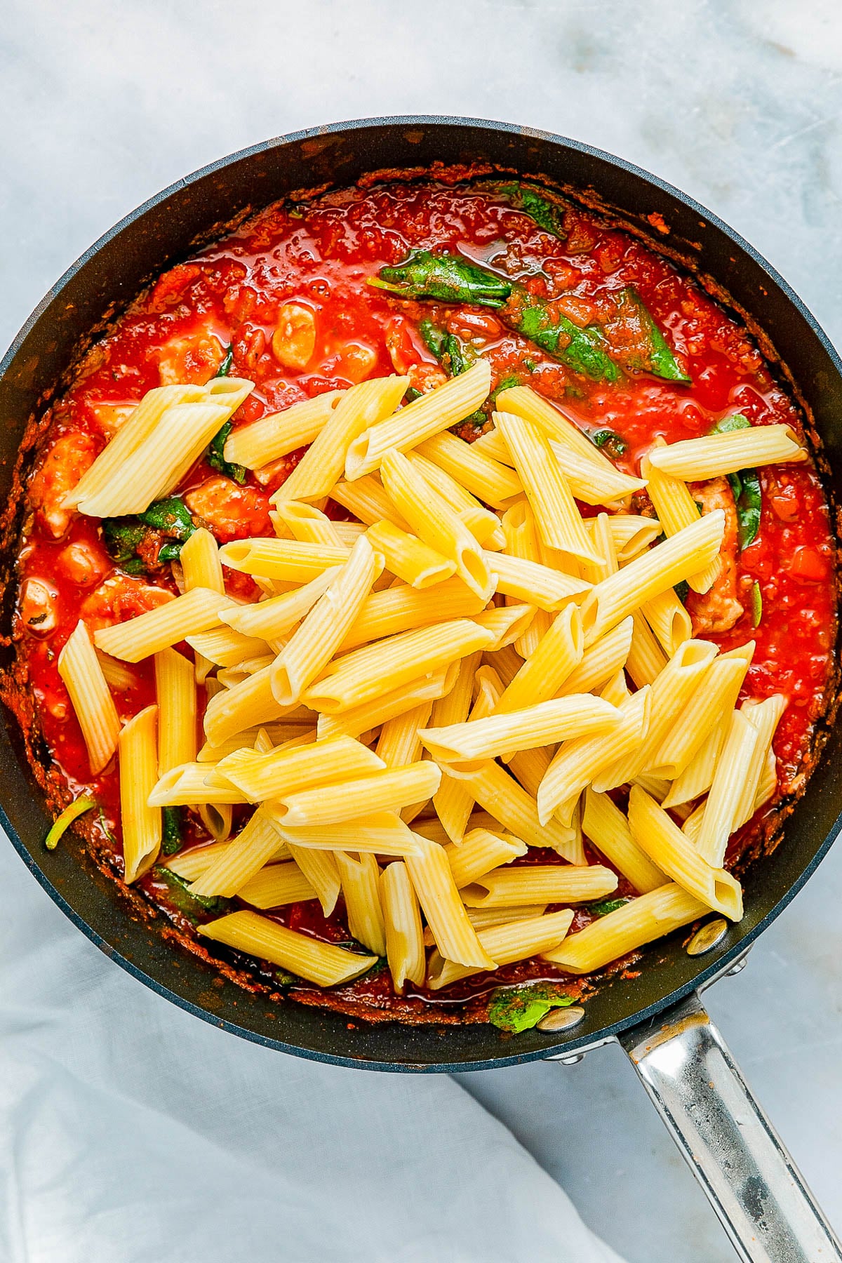 Penne pasta is being mixed into a tomato and spinach sauce in a black skillet.