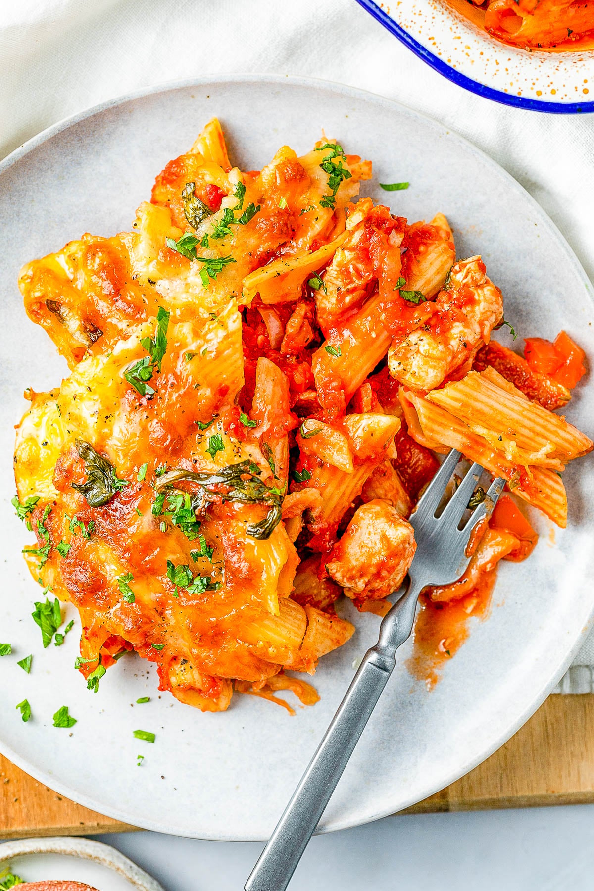 Plate of baked penne pasta with red sauce, cheese, sausage, and herbs. A fork rests on the plate.