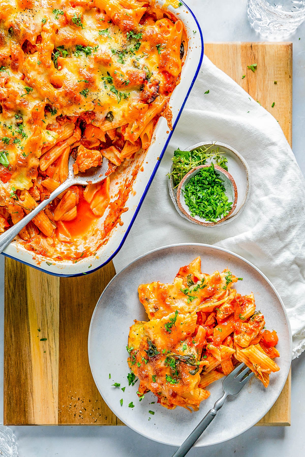 A baked pasta dish with melted cheese is in a white baking dish. A serving is plated on a white plate beside it. A small bowl of chopped parsley is nearby.