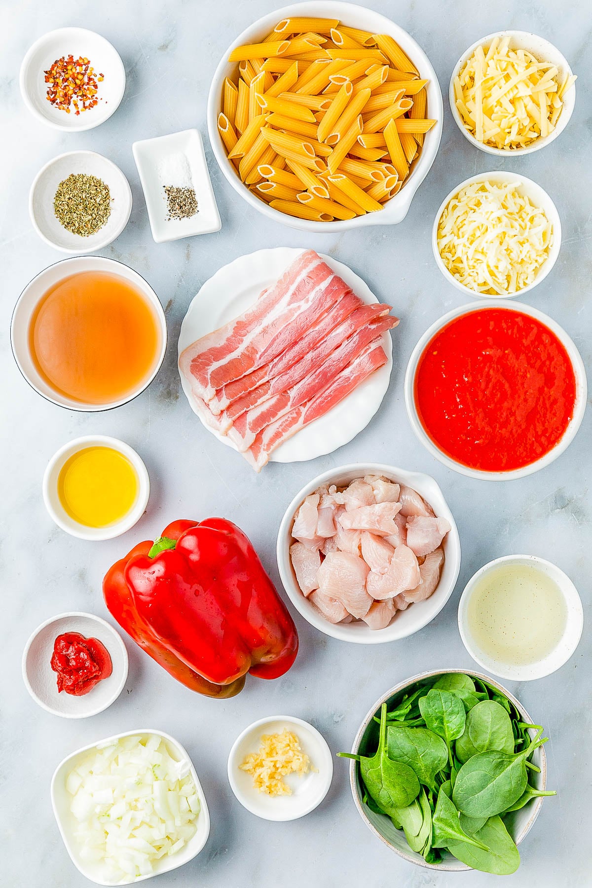A variety of ingredients arranged on a table, including pasta, cheese, raw chicken, bacon, spinach, red pepper, onion, garlic, tomato sauce, and seasonings.