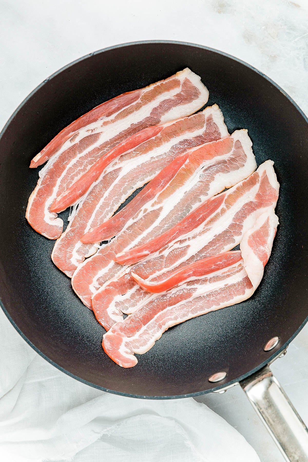 Raw bacon strips in a black frying pan on a white surface.