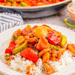 A plate of rice topped with stir-fried chicken, red peppers, and green onions, with a pan of more stir-fry in the background.