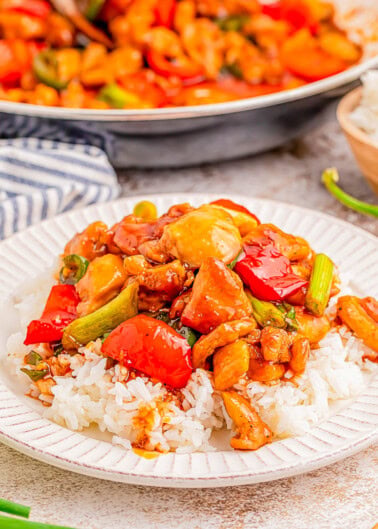 A plate of rice topped with stir-fried chicken, red peppers, and green onions, with a pan of more stir-fry in the background.