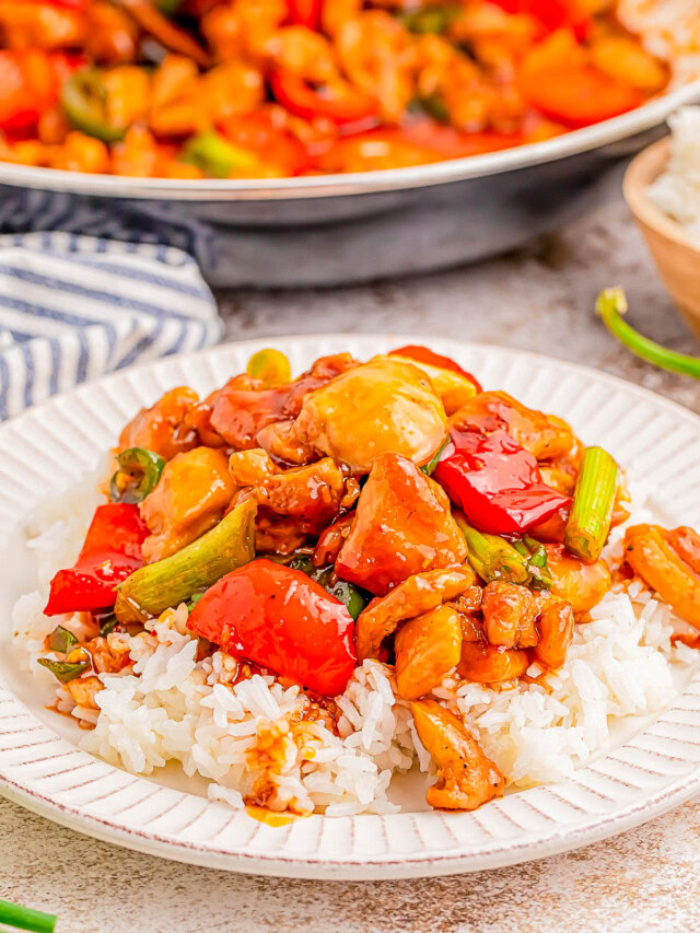 A plate of rice topped with stir-fried chicken, red peppers, and green onions, with a pan of more stir-fry in the background.