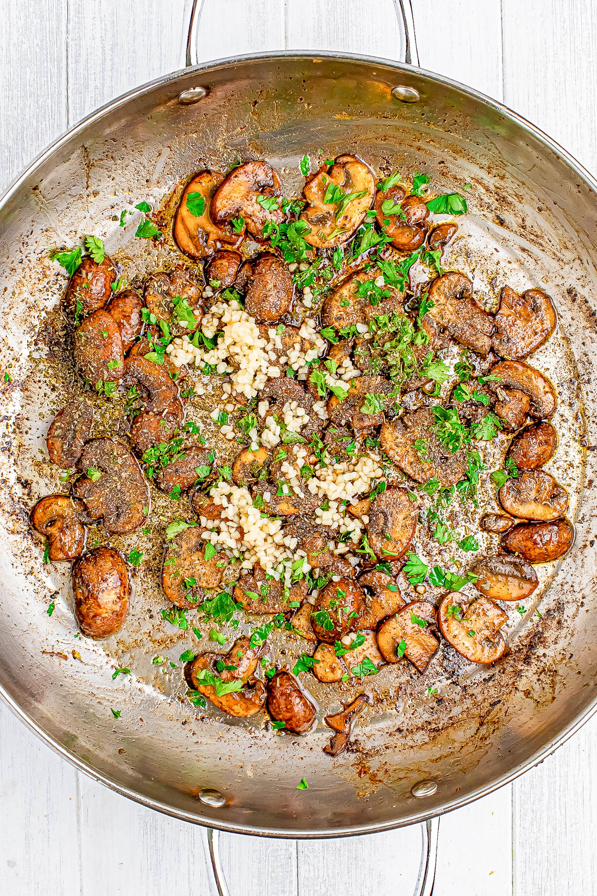 Saut&eacute;ed mushrooms in a skillet with herbs and chopped garlic on a light wooden surface.
