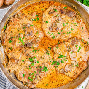 A pan containing cooked pork chops in a creamy mushroom sauce, garnished with chopped parsley. A bowl of mushrooms and parsley are in the background.