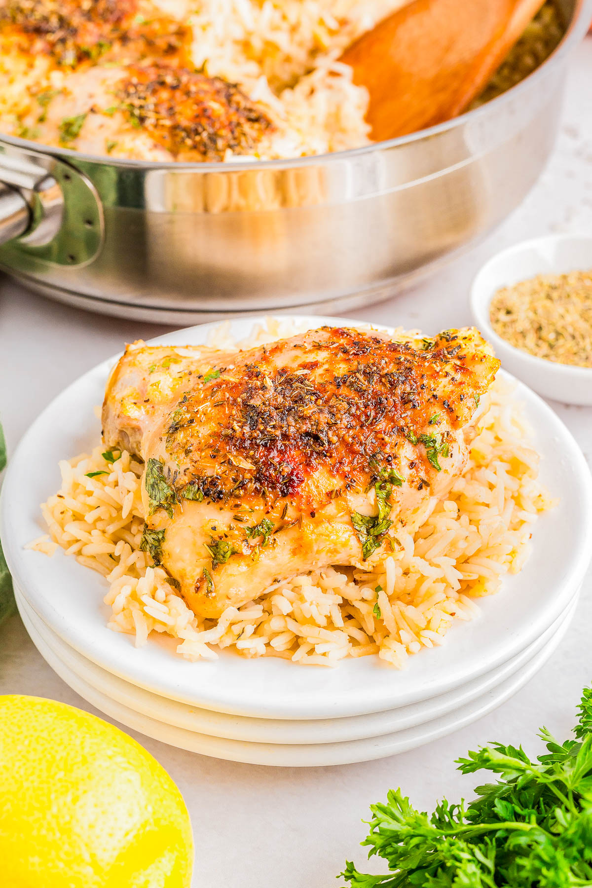 Grilled chicken breast on a bed of rice, garnished with herbs, in a white plate. A pan, small bowl, lemon, and parsley are in the background.