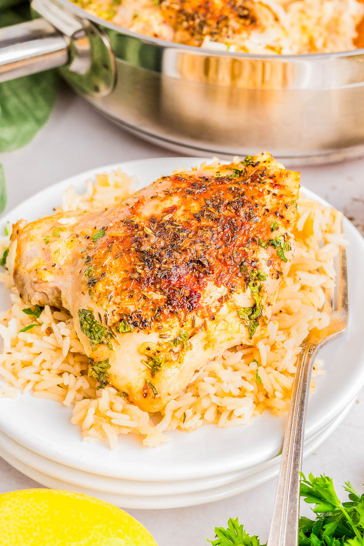 Seasoned chicken breast on a bed of rice, garnished with herbs, served on a white plate with a fork. Skillet and lemon in the background.