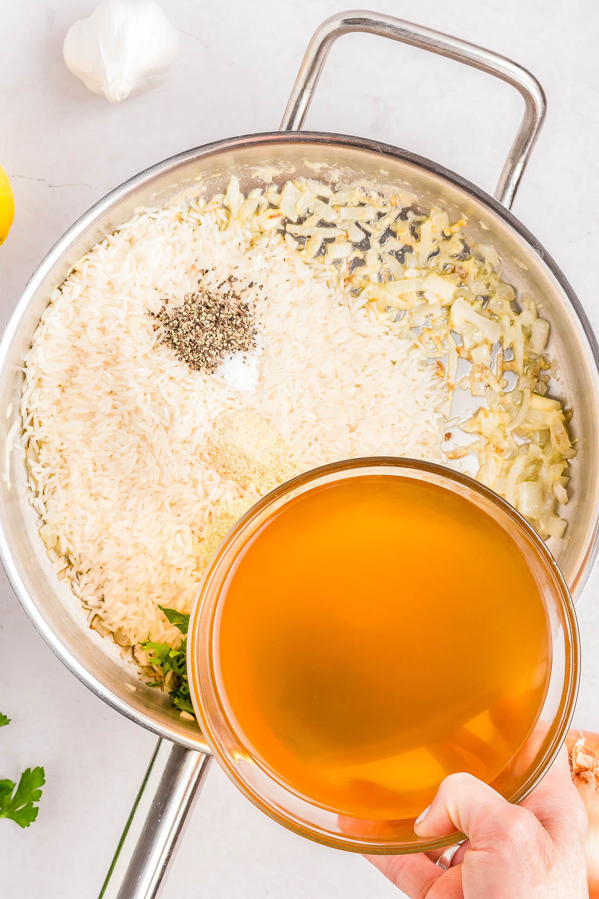 A person pours broth into a pan containing rice, chopped onions, seasoning, and herbs. A garlic clove and half a lemon are visible nearby.