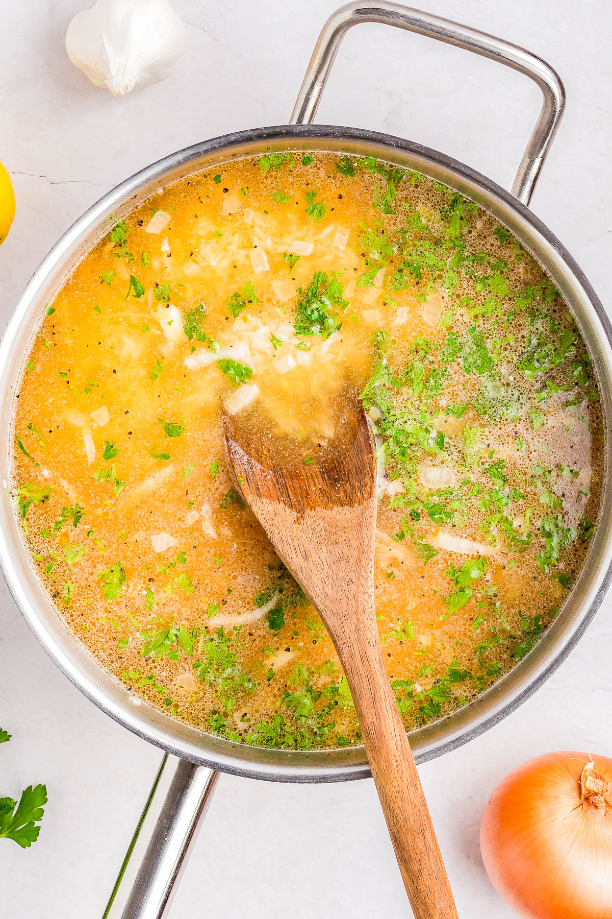 A pot of soup with a wooden spoon, featuring visible herbs, onion pieces, and a light broth. Garlic, lemon, parsley, and an onion are placed around the pot on a white surface.