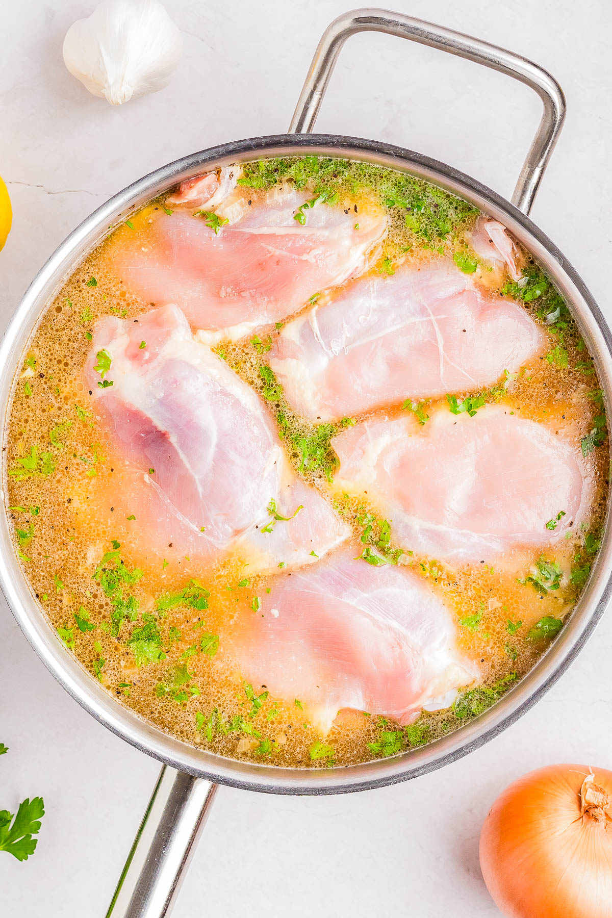 Raw chicken breasts marinating in a metal pan with a liquid containing herbs and spices, surrounded by garlic, lemon, and onion on a white surface.