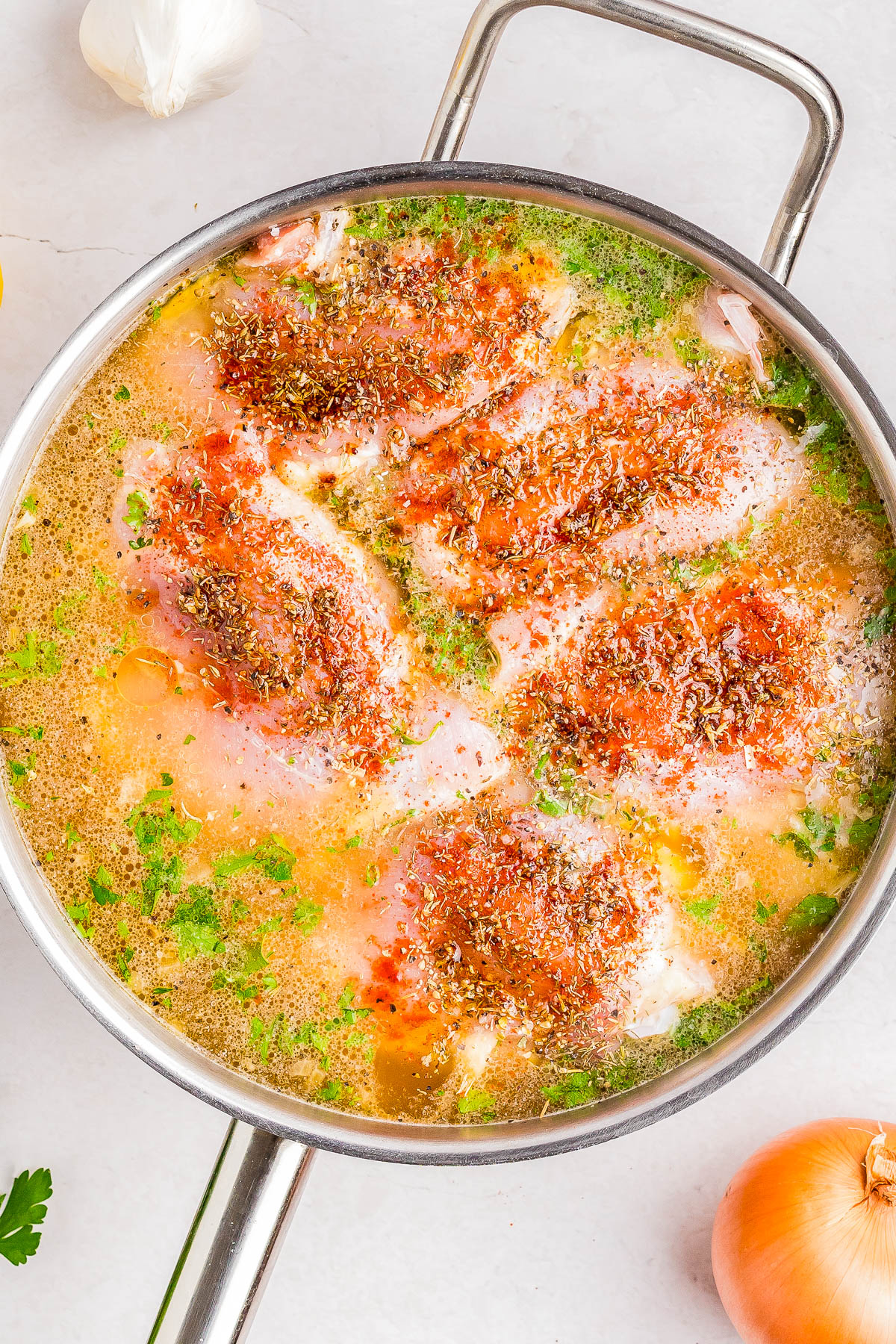 A pot with raw chicken breasts seasoned with spices and herbs in a liquid broth, surrounded by garlic and an onion on a light countertop.
