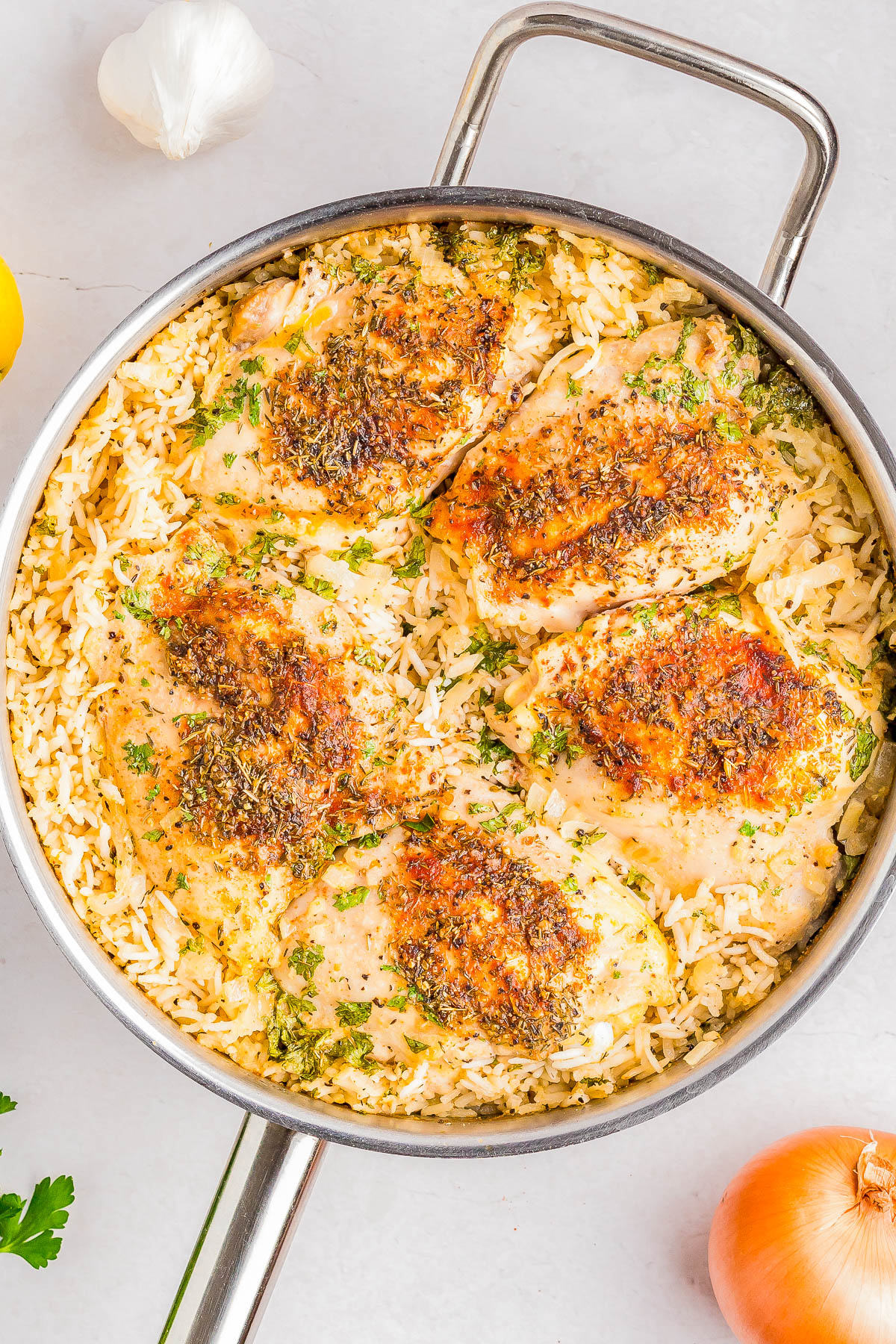 A skillet with cooked, seasoned chicken breasts on a bed of rice, garnished with herbs. Nearby are a garlic bulb, lemon, onion, and parsley on a light surface.