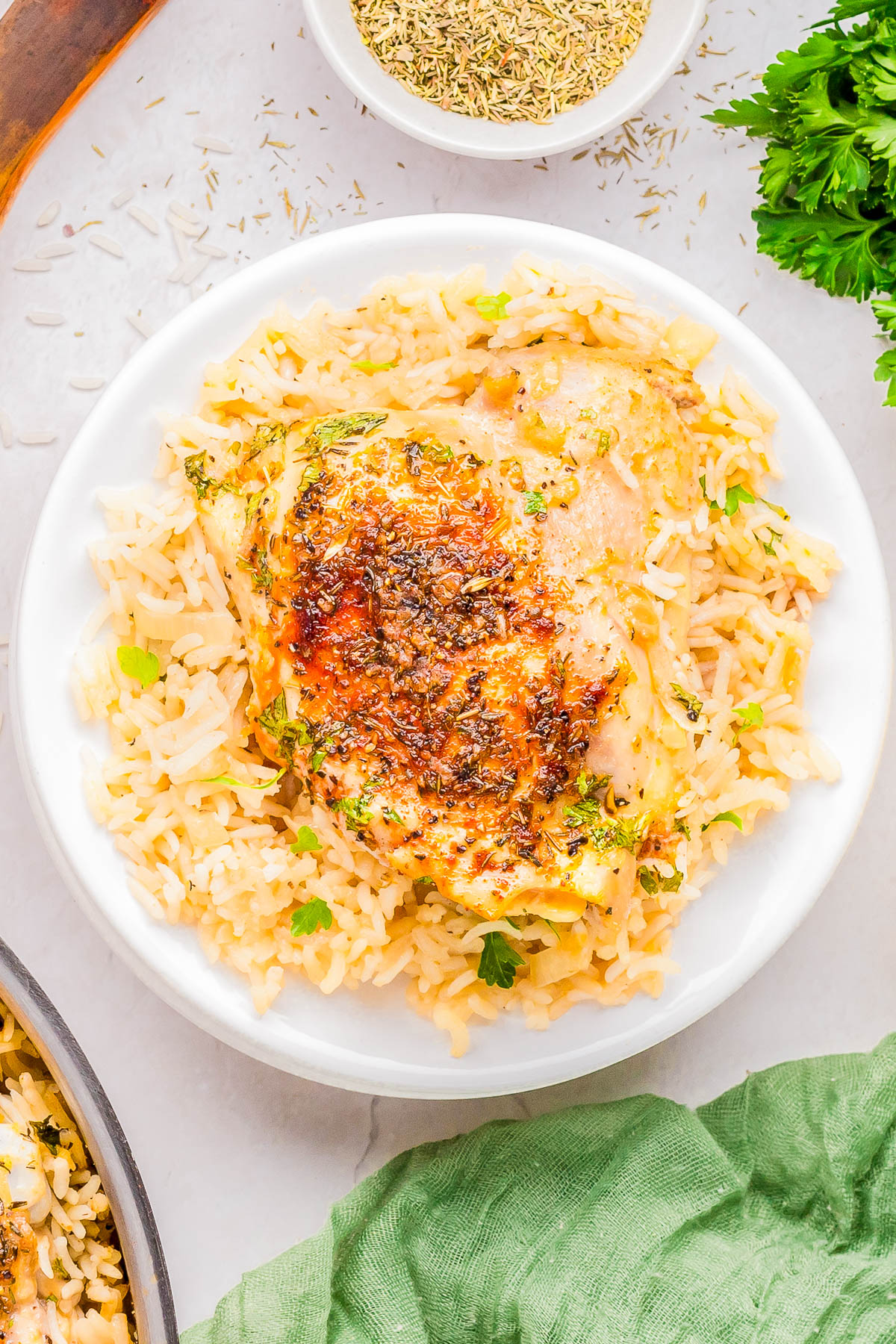 A plate of seasoned chicken breast on a bed of rice, garnished with fresh herbs, surrounded by a small bowl of dried herbs and parsley on a light surface.