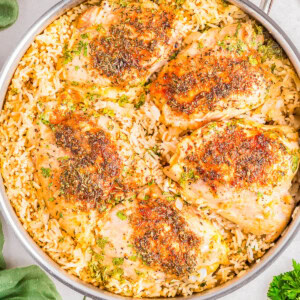 A skillet with seasoned chicken thighs on a bed of cooked rice, garnished with herbs.