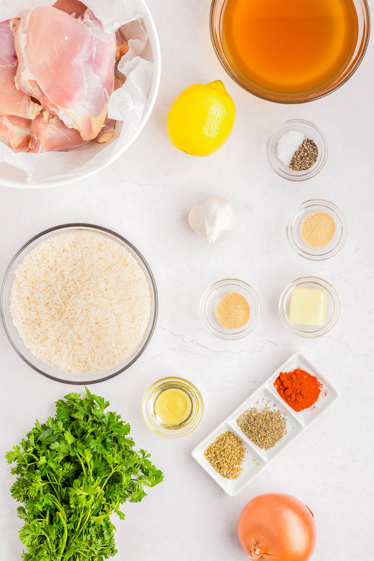Ingredients for a recipe displayed: chicken, lemon, broth, rice, garlic, onion, spices, herbs, butter, oil, salt, and pepper on a white surface.
