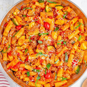 A pan of rigatoni pasta with ground meat, zucchini, cherry tomatoes, and grated cheese, garnished with fresh basil.