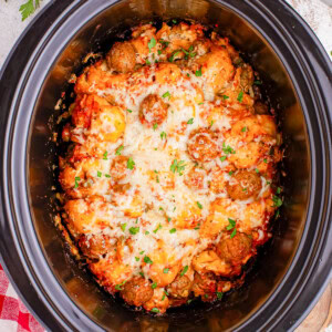 A slow cooker containing meatballs in a tomato sauce, topped with melted cheese and garnished with chopped herbs.