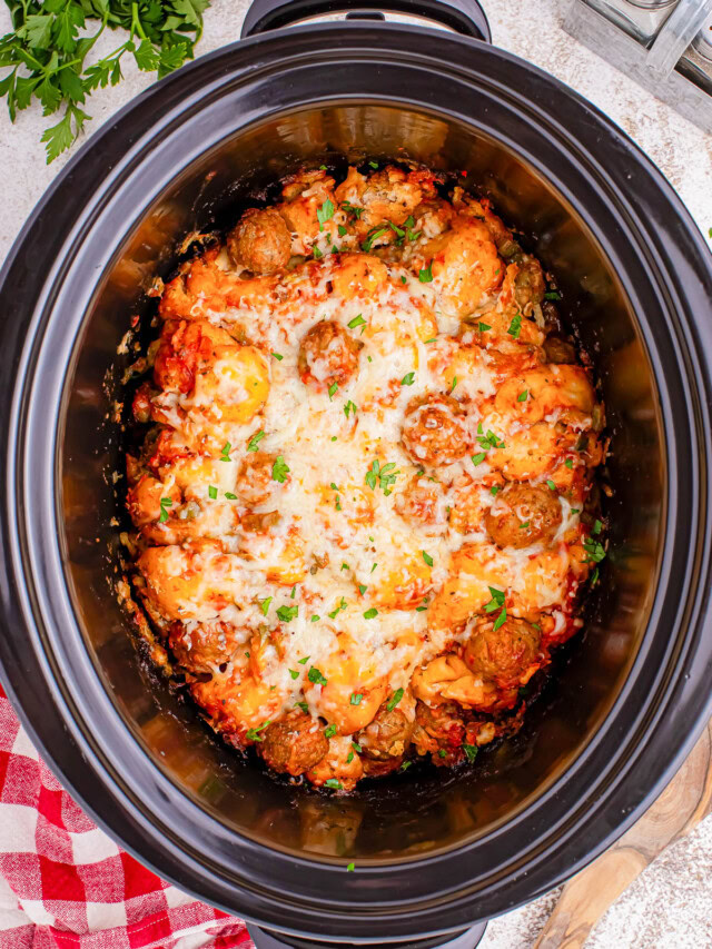 A slow cooker containing meatballs in a tomato sauce, topped with melted cheese and garnished with chopped herbs.