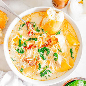 Bowl of creamy soup with spinach, shredded cheese, lasagna noodles, and chicken, garnished with sun-dried tomatoes and served with a piece of bread. Crockpot and pepper mill in the background.