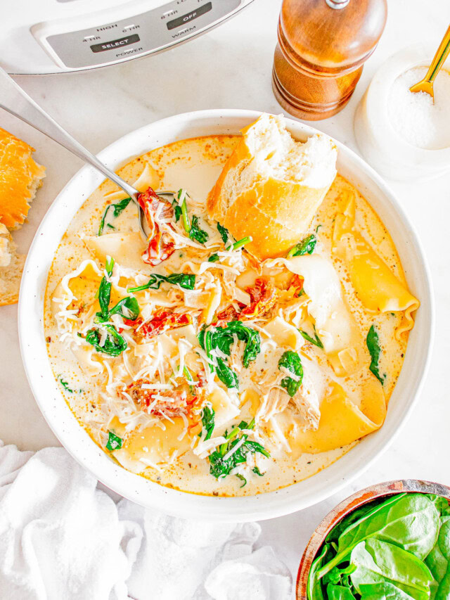 Bowl of creamy soup with spinach, shredded cheese, lasagna noodles, and chicken, garnished with sun-dried tomatoes and served with a piece of bread. Crockpot and pepper mill in the background.