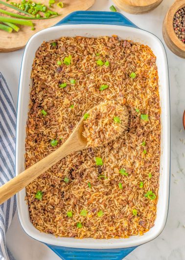 A casserole dish filled with baked rice topped with green onions. A wooden spoon rests in the dish. Surrounding items include chopped green onions, pepper, salt, and a striped cloth.