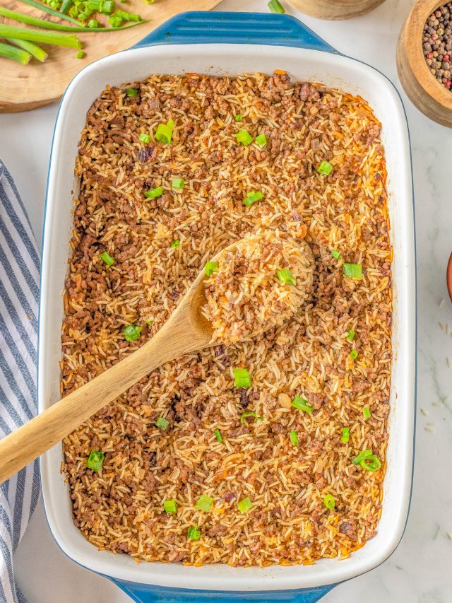 A casserole dish filled with baked rice topped with green onions. A wooden spoon rests in the dish. Surrounding items include chopped green onions, pepper, salt, and a striped cloth.