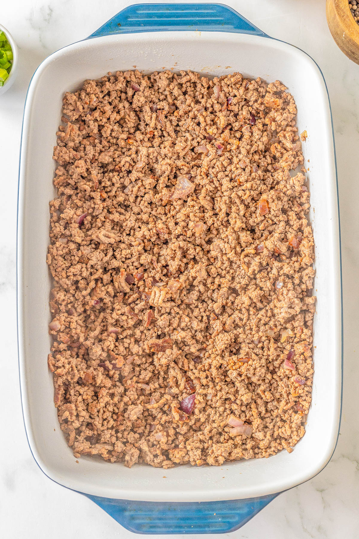 Ground meat with diced onions in a rectangular blue and white baking dish on a marble surface.