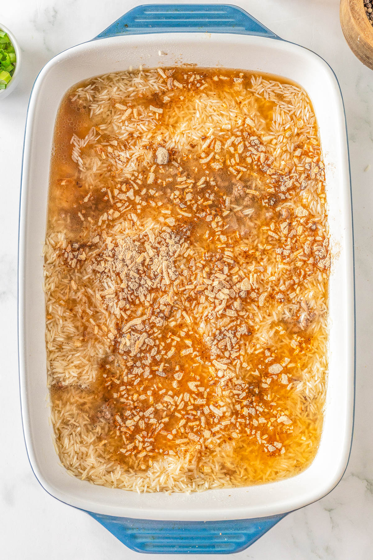 Uncooked rice mixed with soup and seasonings in a blue-rimmed rectangular baking dish.