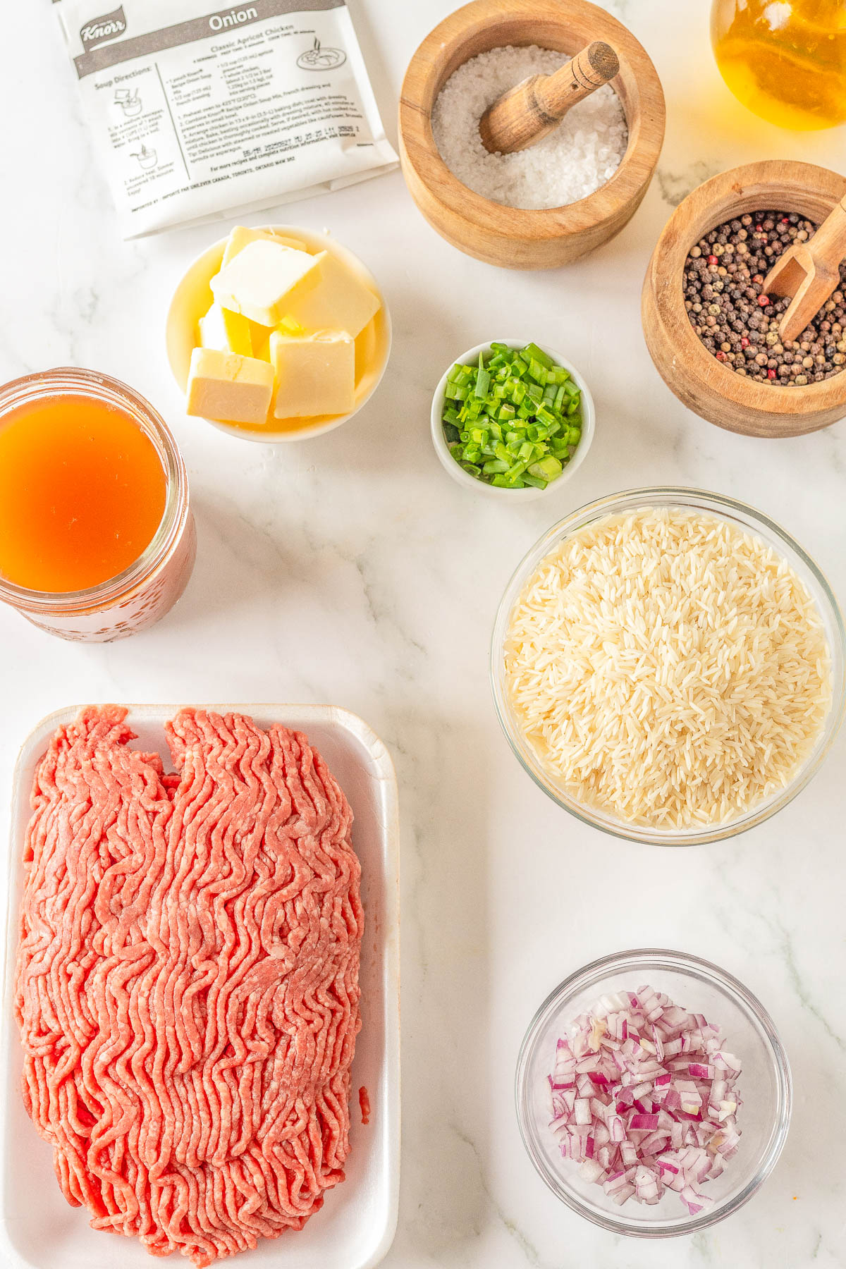 Ingredients on a marble countertop: ground meat, rice, butter cubes, chopped onions, green onions, beef broth, salt, pepper, and a packet labeled