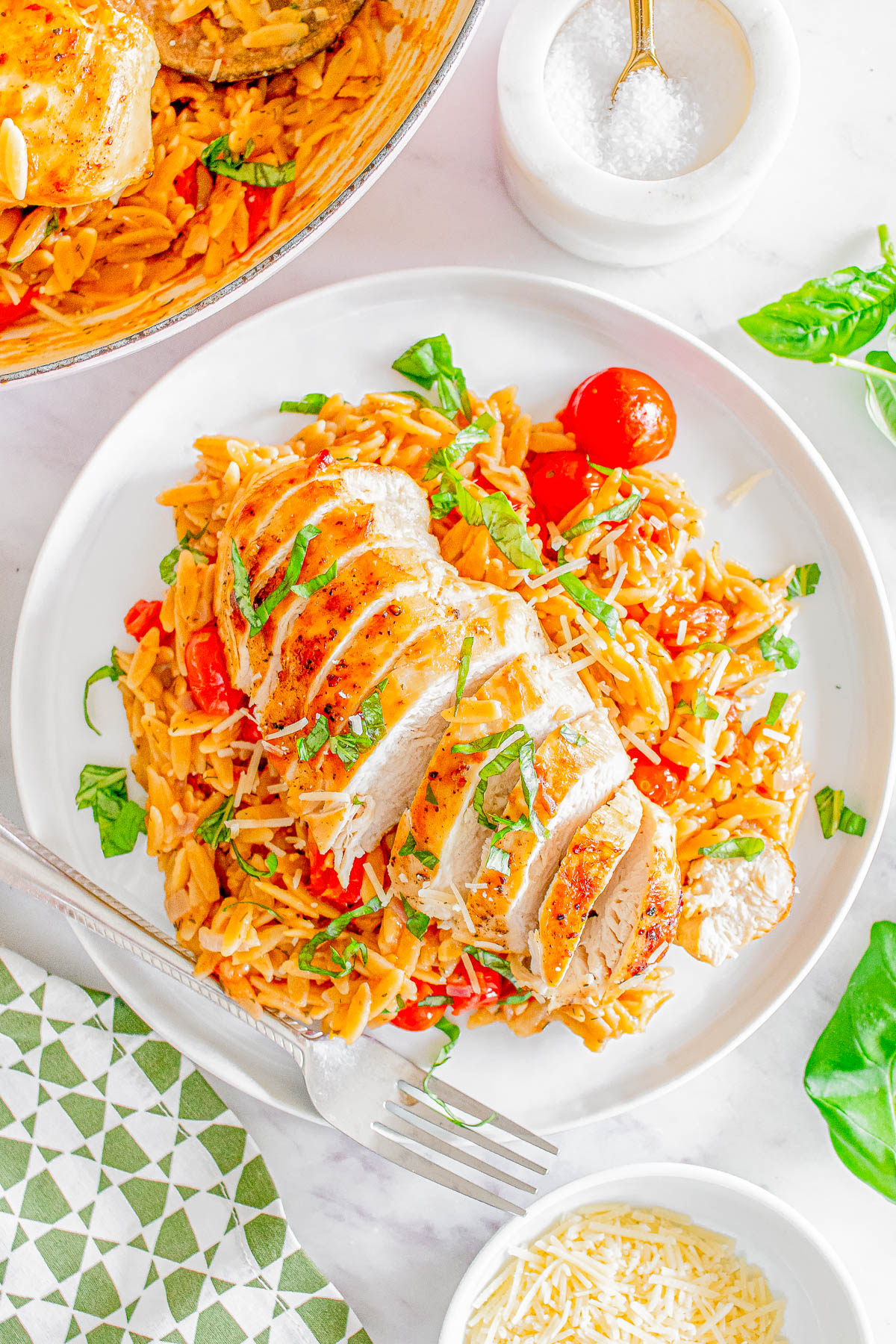A plate with sliced chicken breast on a bed of orzo with cherry tomatoes and herbs. A fork and knife are placed beside the plate.