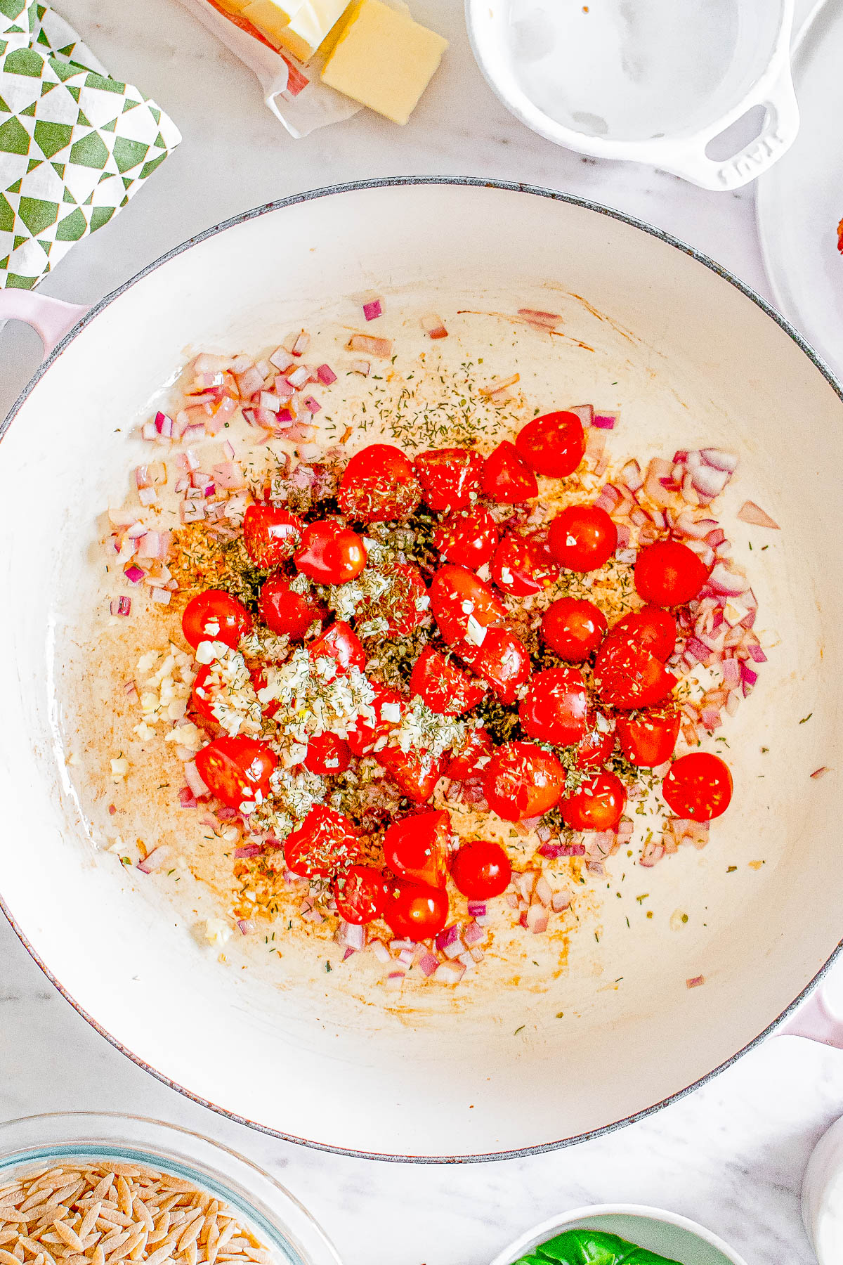 Chopped tomatoes, onions, and spices in a white skillet, with ingredients like butter and herbs nearby on a marble surface.