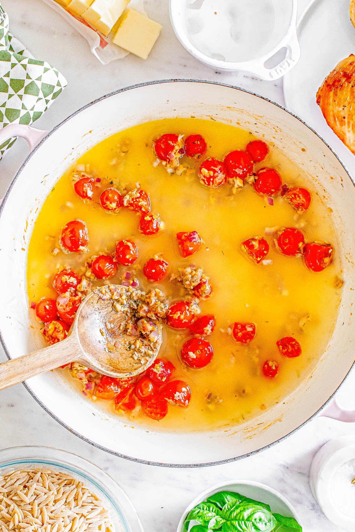 A pot on a stove contains cherry tomatoes and ground sausage in a yellow oil-based sauce. A wooden spoon rests on the mixture. There are ingredients and kitchen items around the pot.
