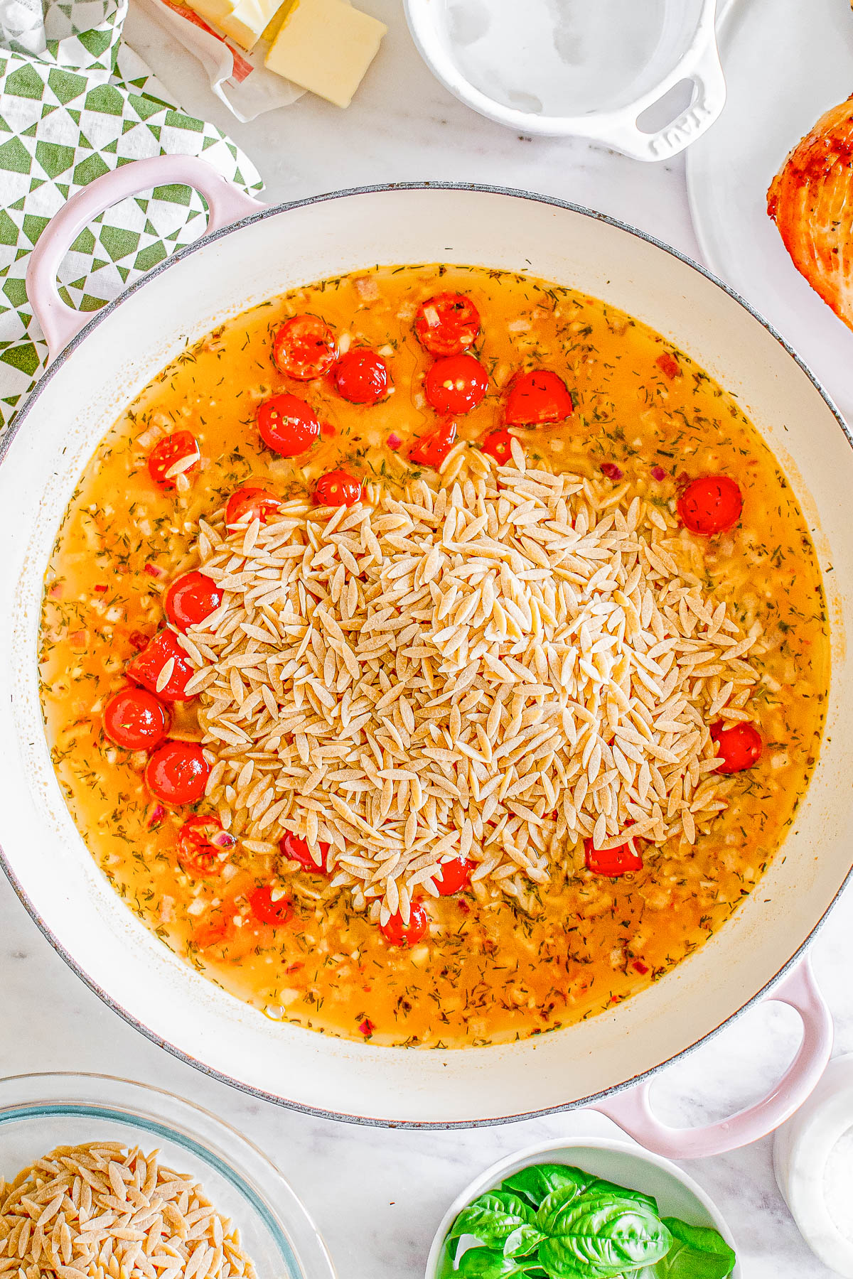 Pot filled with uncooked orzo, cherry tomatoes, herbs, and broth, ready for cooking.