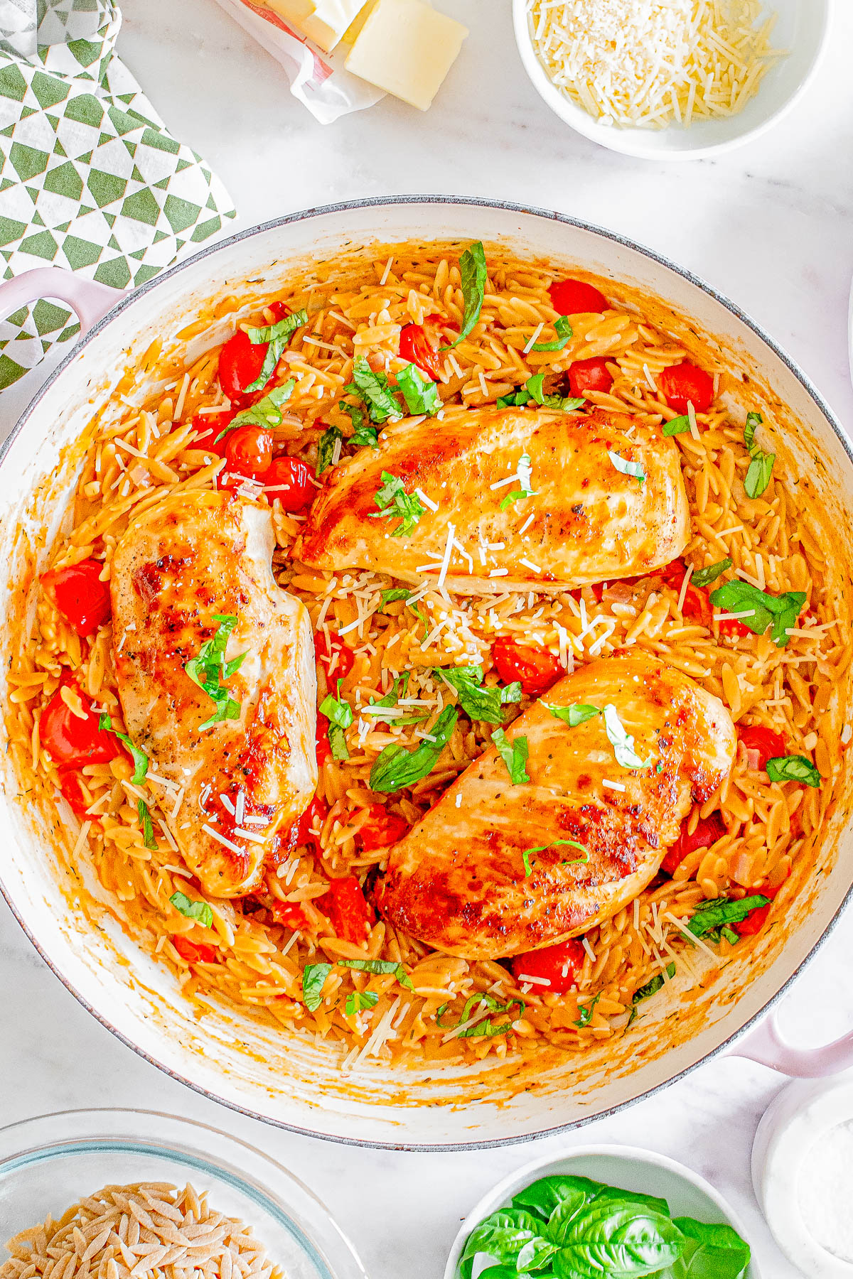 Skillet with cooked chicken breasts on a bed of creamy orzo, red bell peppers, topped with grated cheese and fresh basil leaves.