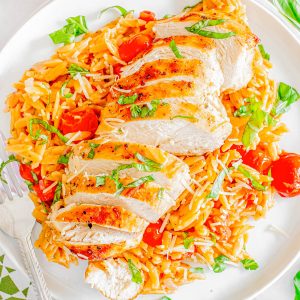 Sliced grilled chicken served on a bed of orzo pasta with cherry tomatoes and topped with grated cheese and basil on a white plate.