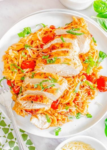 Sliced grilled chicken served on a bed of orzo pasta with cherry tomatoes and topped with grated cheese and basil on a white plate.
