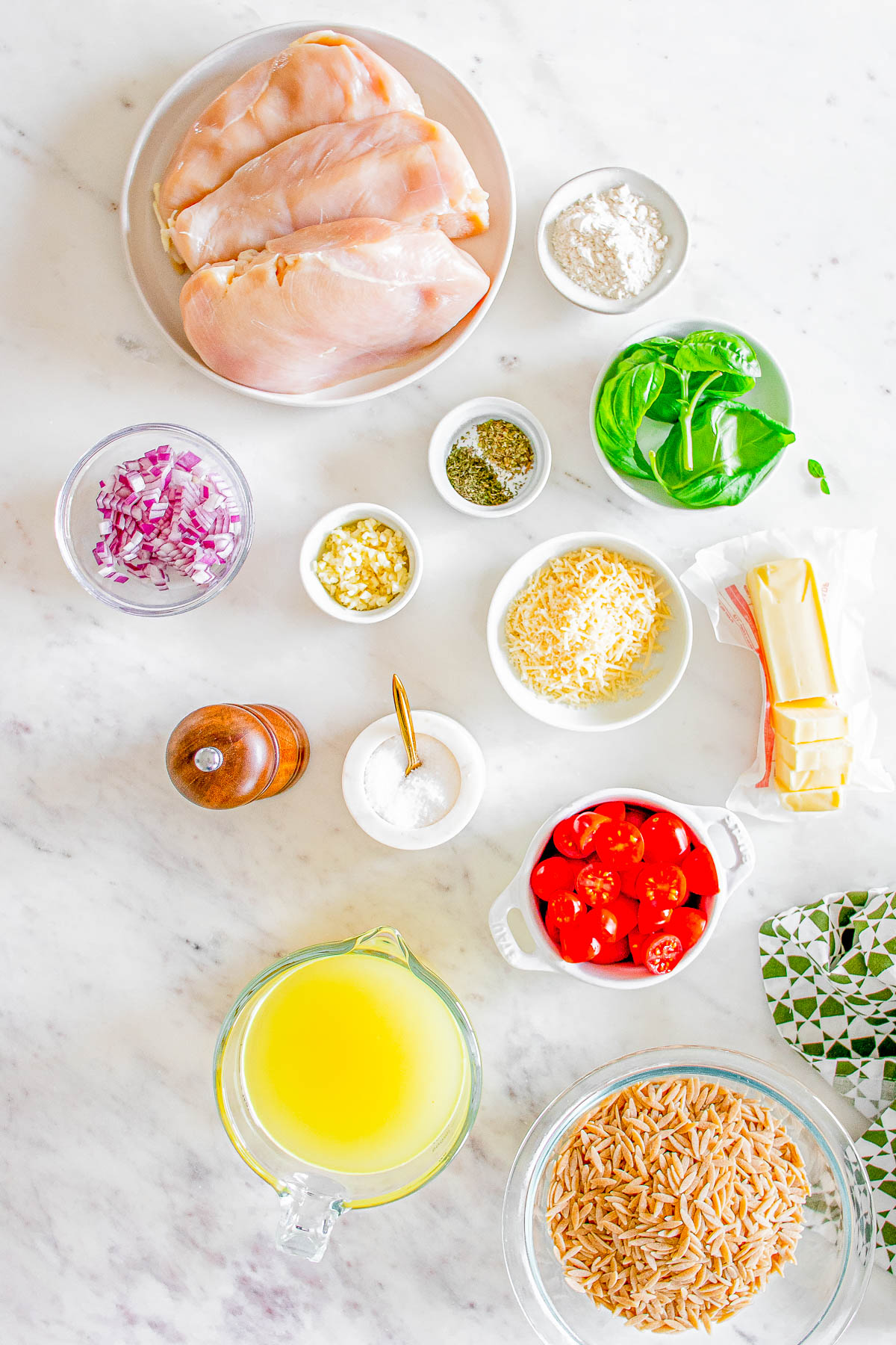 Ingredients for a dish arranged on a countertop: raw chicken breasts, diced red onion, garlic, cherry tomatoes, spices, cheese, broth, pasta, and butter.