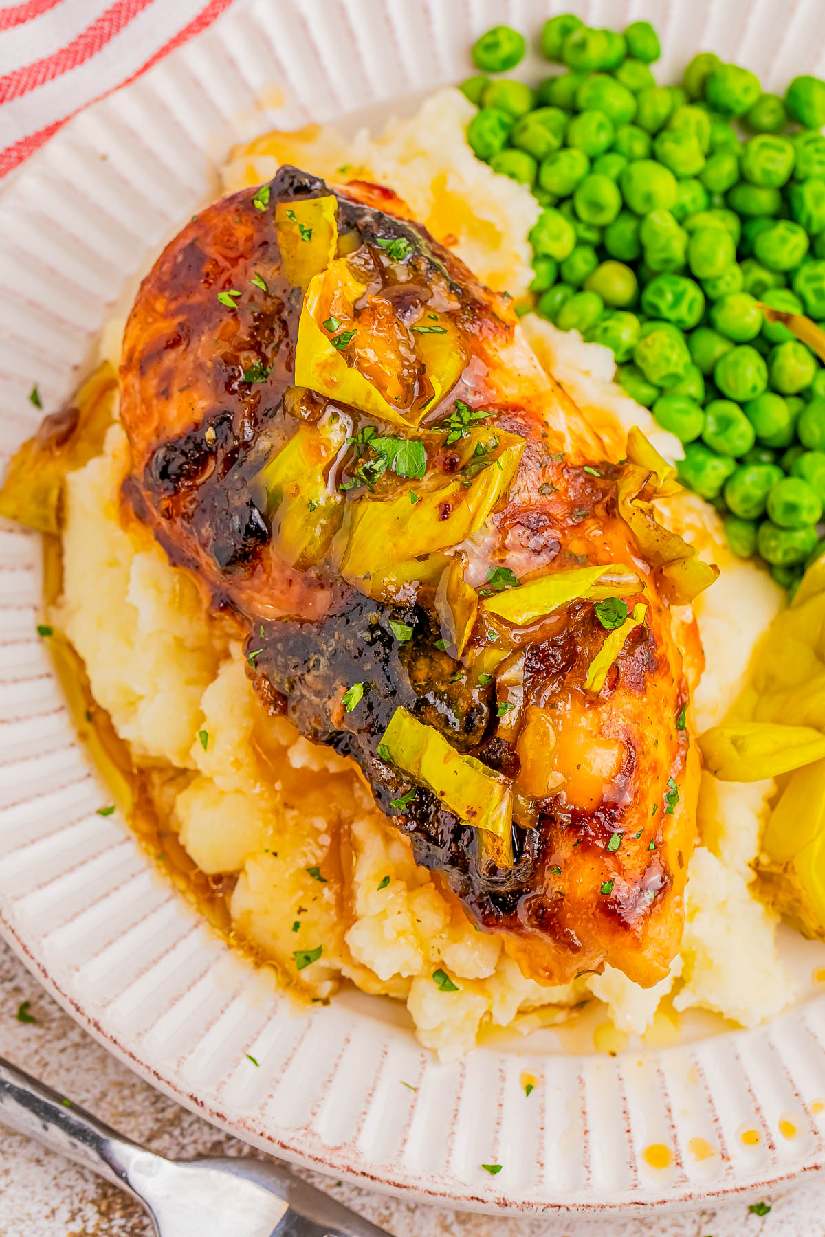 Grilled chicken topped with sauce and herbs on mashed potatoes, served with green peas and sliced vegetables on a white plate.