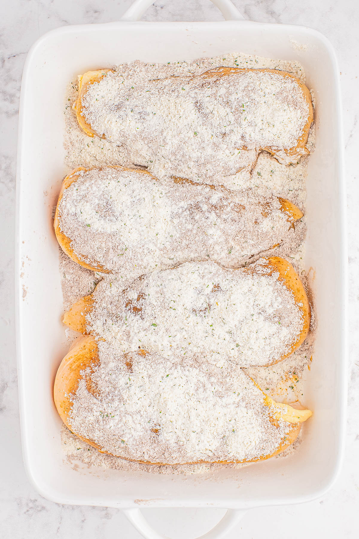 Four slices of chicken coated with a layer of powdered mixture in a white baking dish.