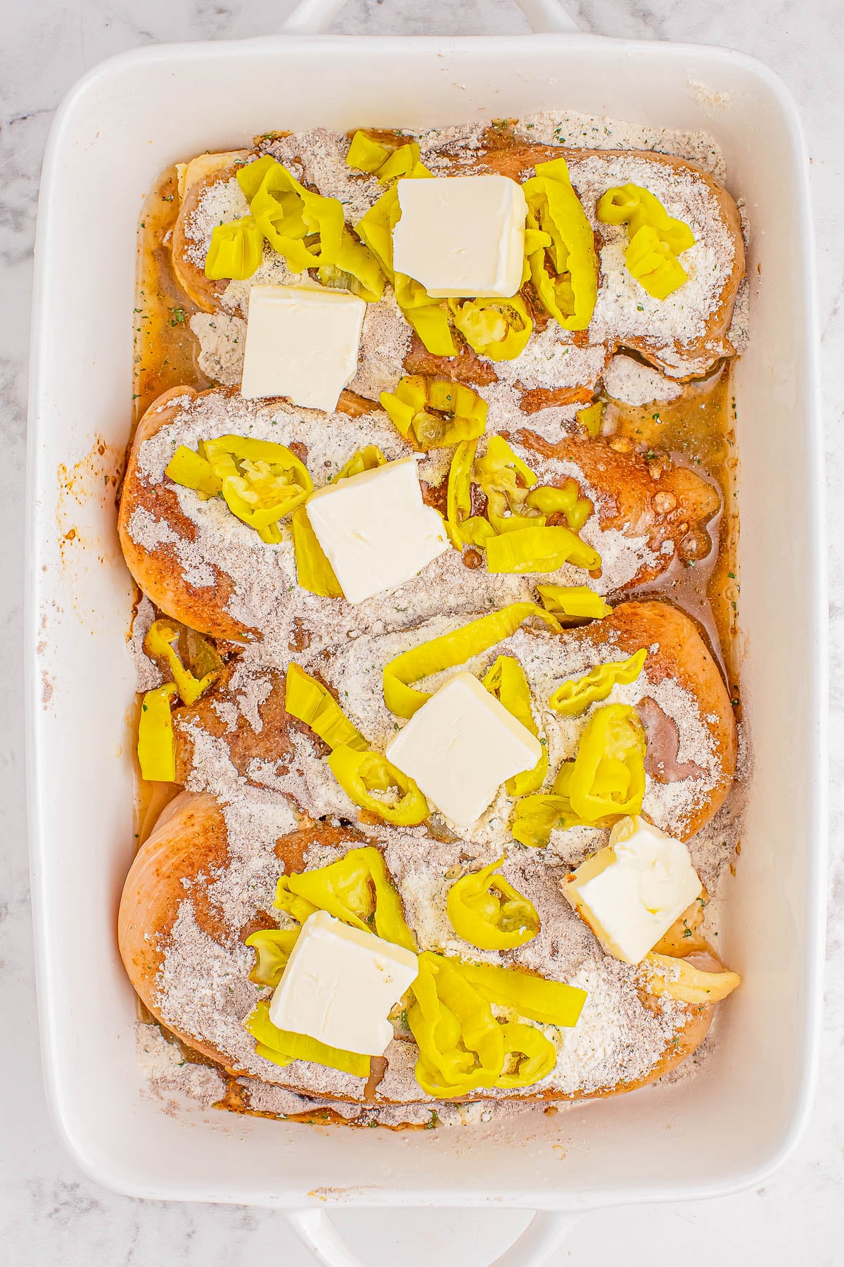 Raw chicken breasts in a white baking dish topped with seasoning, pats of butter, and sliced yellow peppers.