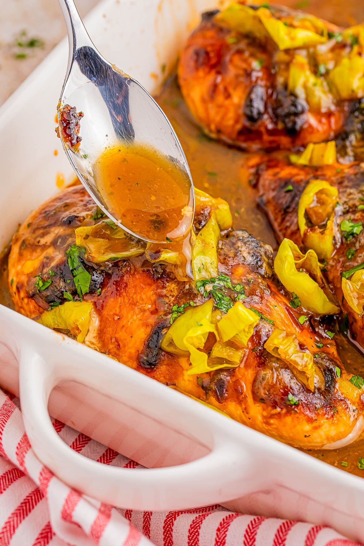 A spoon drizzling sauce over cooked chicken breasts topped with sliced peppers in a white baking dish.