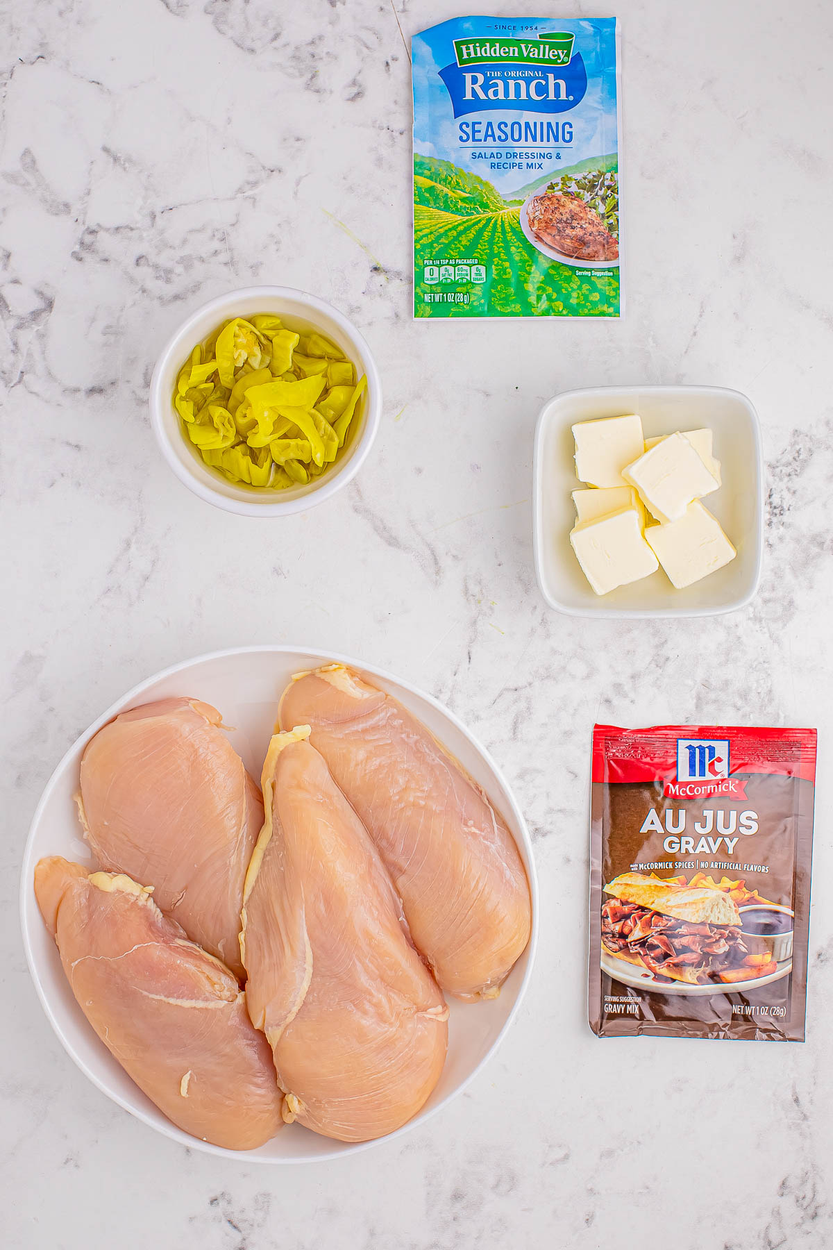 Ingredients on a countertop: raw chicken breasts on a plate, a bowl of sliced peppers, a bowl of butter cubes, a packet of ranch seasoning, and a packet of au jus gravy mix.