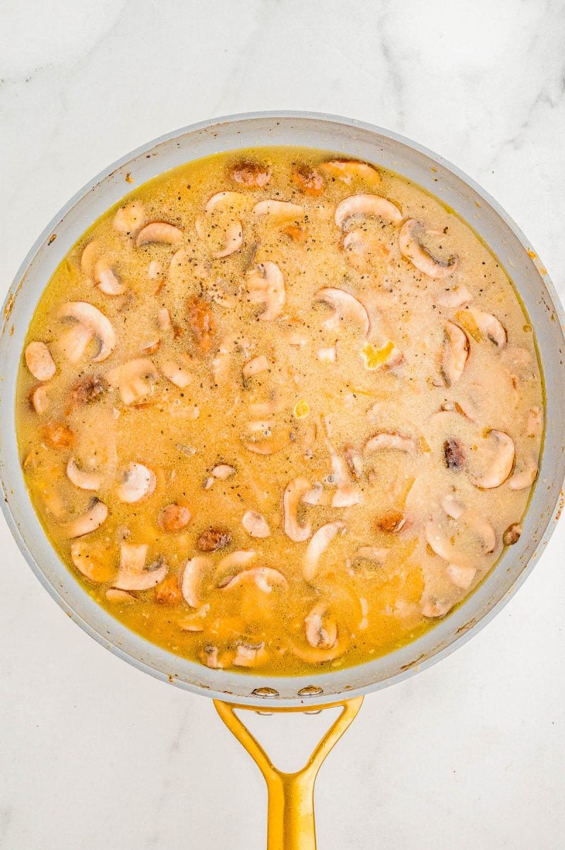 A frying pan filled with creamy mushroom sauce, featuring sliced mushrooms in a light brown liquid, resting on a marble surface.