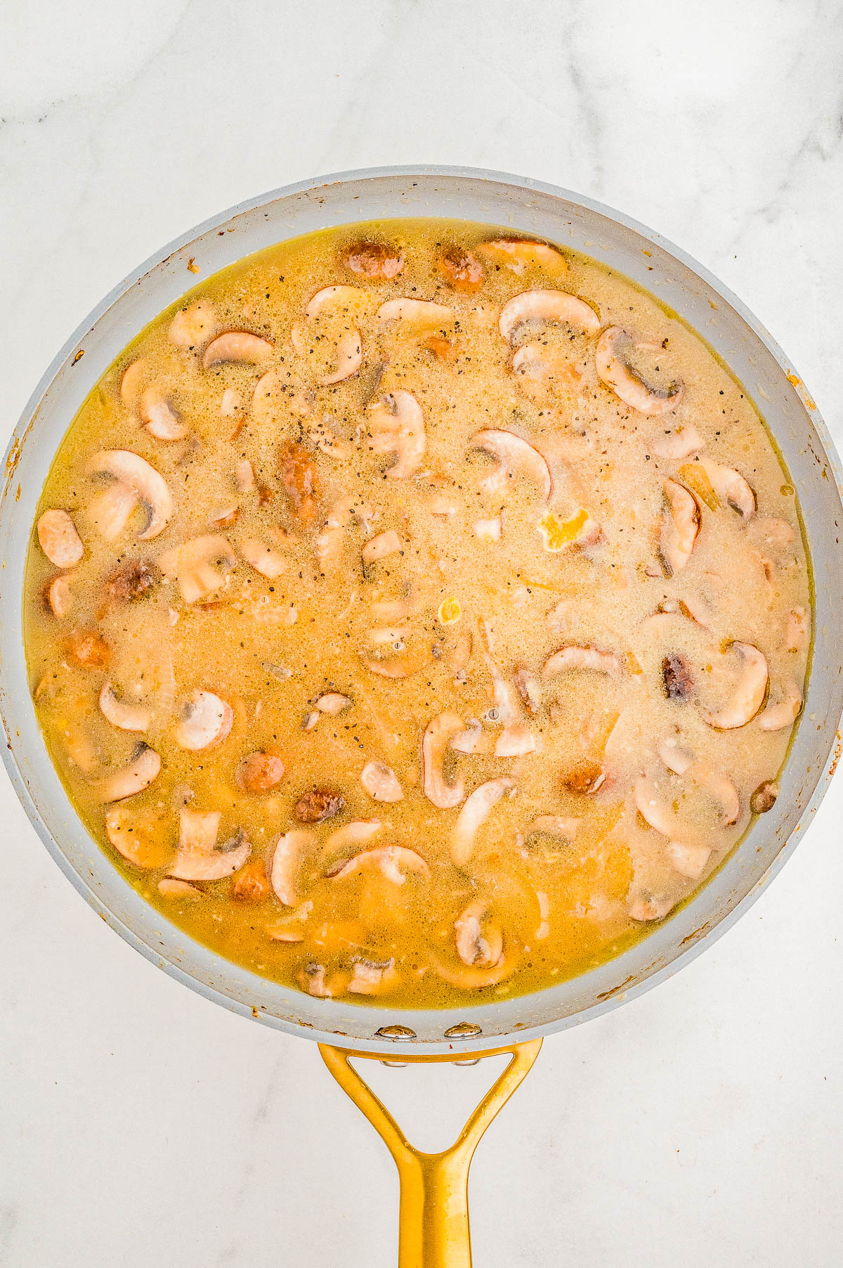 A frying pan filled with creamy mushroom sauce, featuring sliced mushrooms in a light brown liquid, resting on a marble surface.