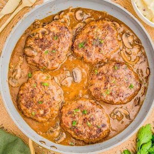 Five beef patties in a mushroom gravy sauce in a skillet, garnished with parsley, next to mashed potatoes and fresh herbs.