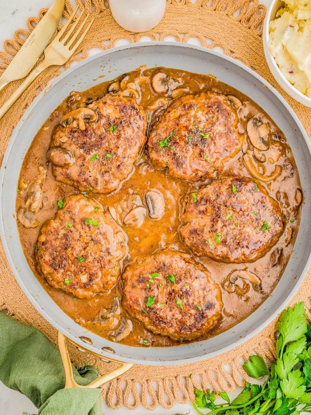 Five beef patties in a mushroom gravy sauce in a skillet, garnished with parsley, next to mashed potatoes and fresh herbs.