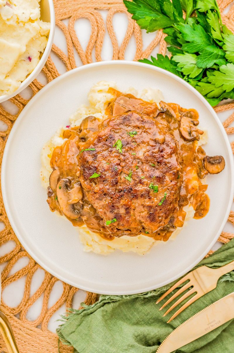 A hamburger steak with mushroom gravy on mashed potatoes, garnished with parsley, is served on a white plate. Accompanied by a fork, knife, a green napkin, and a bowl of mashed potatoes.